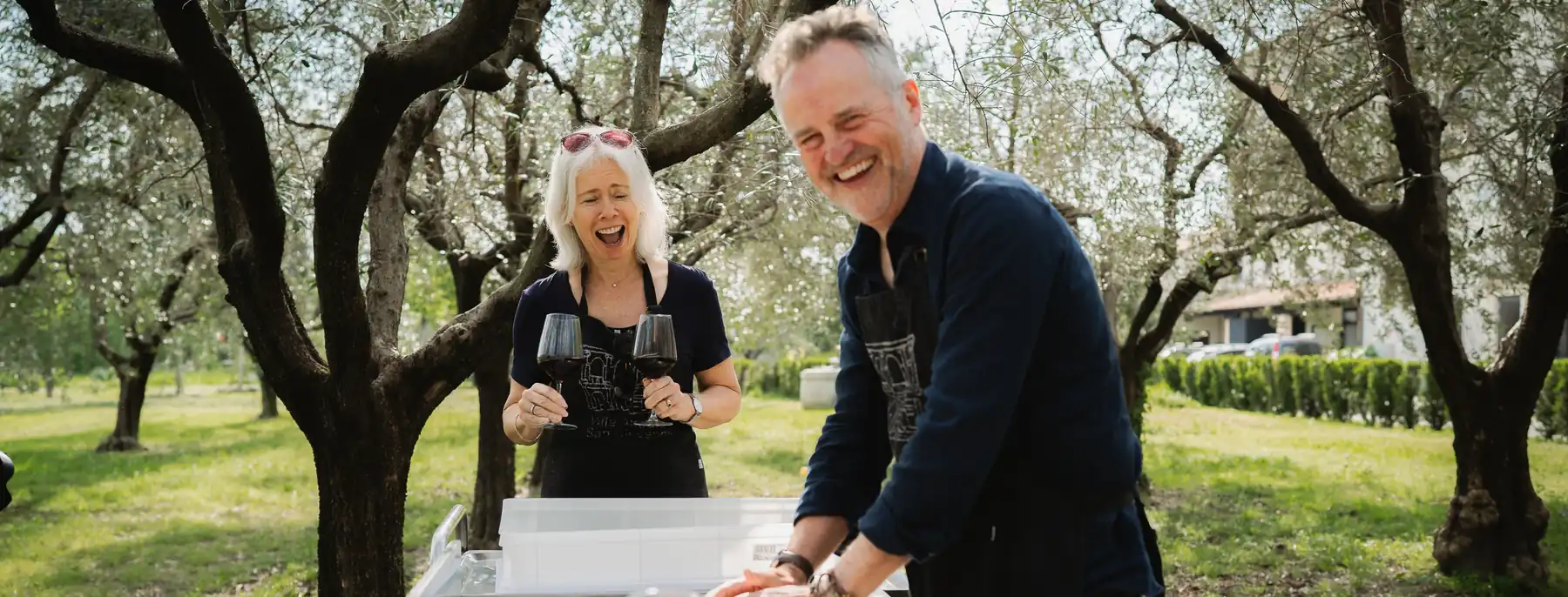 Two guests laughing with glasses of red wine at a table among the olive trees