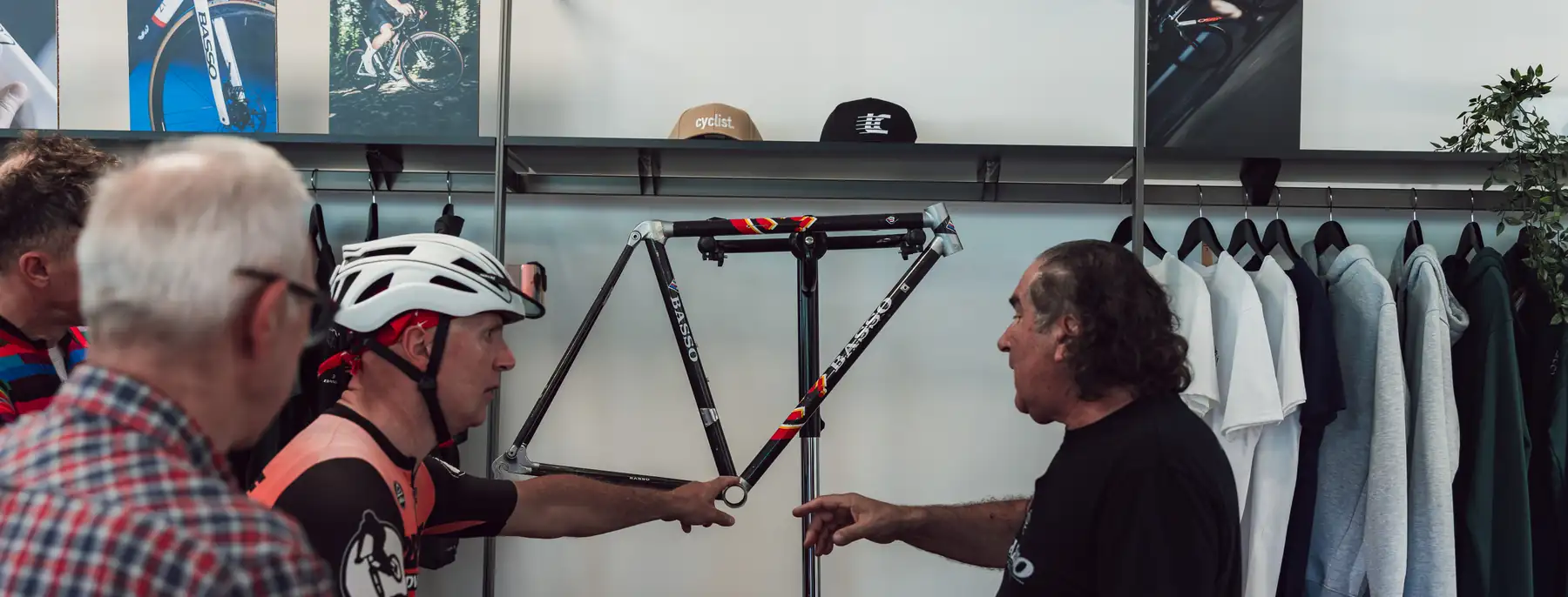 A man in a Basso T-shirt pointing at a bare Basso frame as he explains it to visiting riders in the showroom