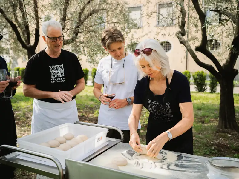 Guests in aprons shaping focaccia dough at an outdoor table under olive trees, glasses of wine in hand