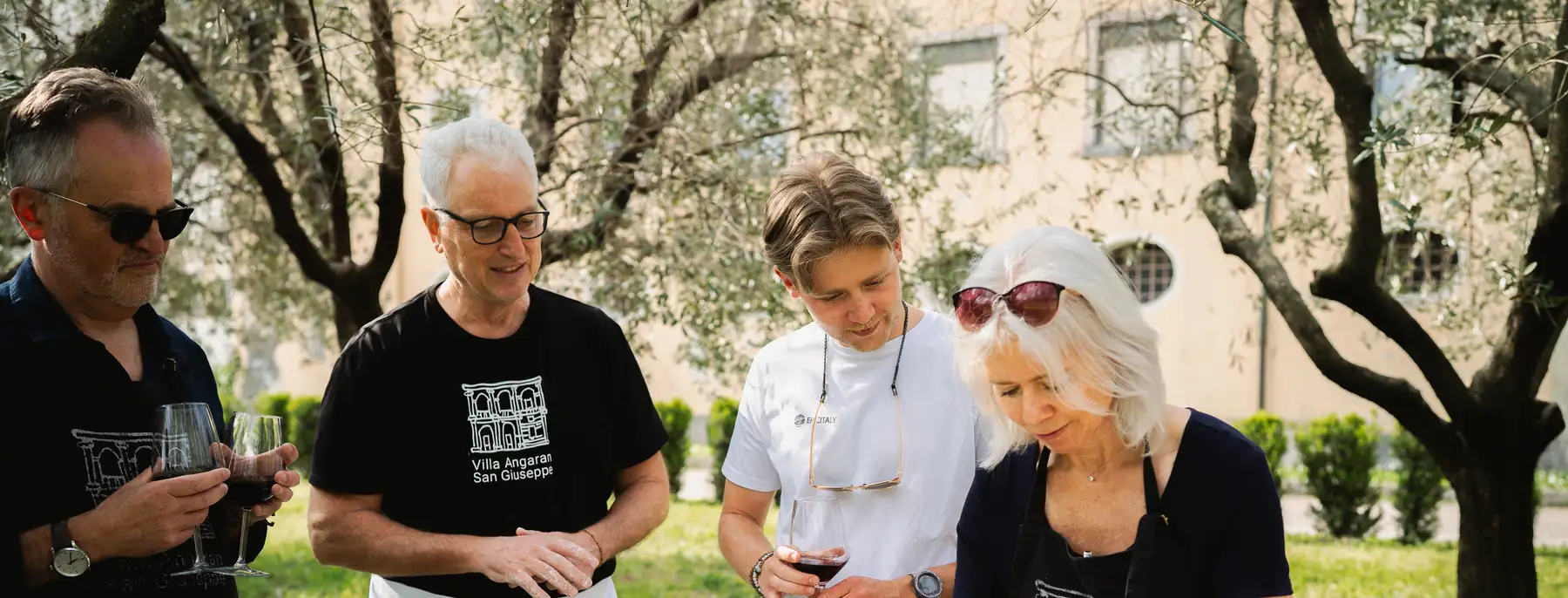 Guests in aprons shaping focaccia dough at an outdoor table under olive trees, glasses of wine in hand