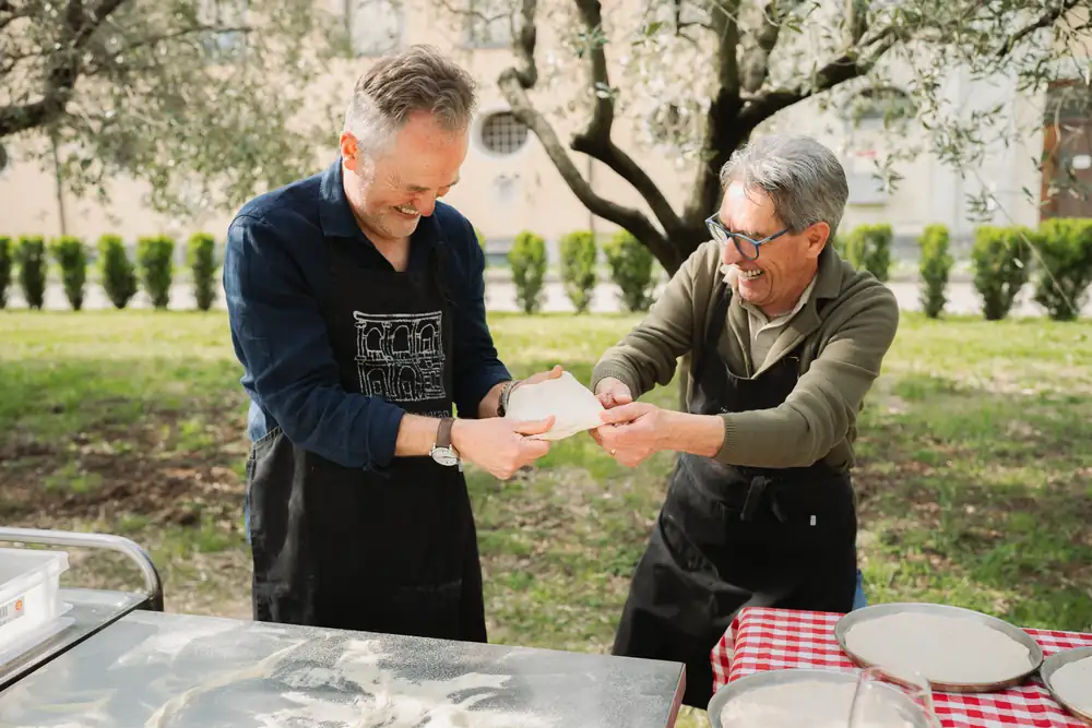 Two men in aprons stretching dough at an outdoor table under the olive trees