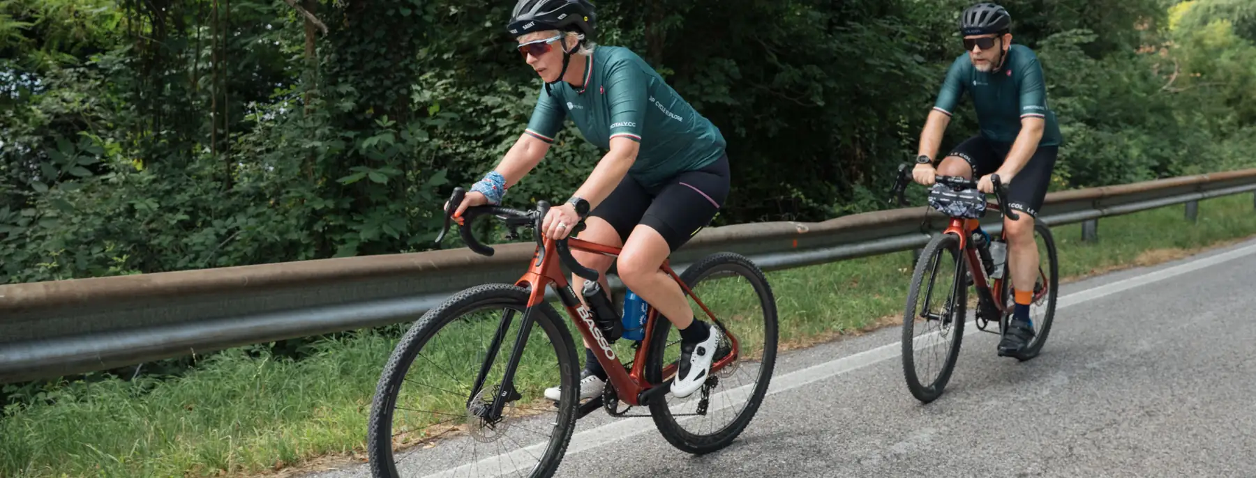 Two riders in matching kit on a quiet country road, with woodland behind